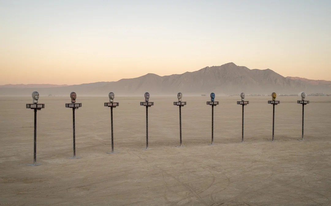 The finished installation on the open playa at dusk — nine heads on tall poles in a row, mountains behind.